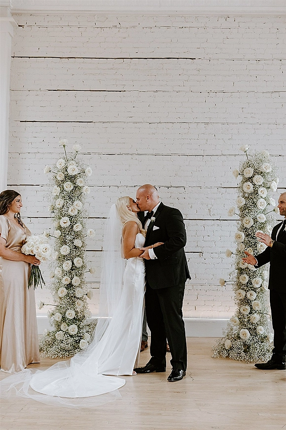 Wedding kiss as bride in satin dress and long veil kisses groom in black tuxedo before white rose floral pillars and brick wall