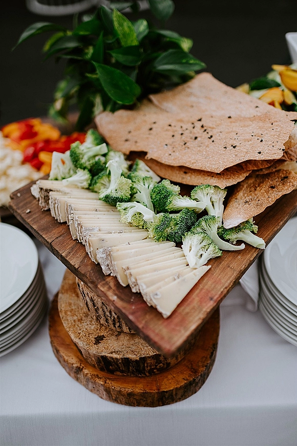 Wedding cheese board with wedding cocktail hour food, featuring cheese slices, crackers, broccoli florets, and greenery on a wooden board at table setting