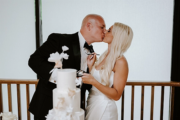 Wedding cake cutting as bride in a strapless gown and groom in a black tuxedo with bow tie share cake forks by window light