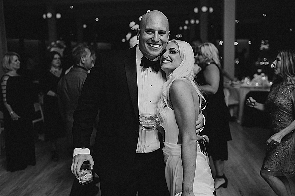 Wedding reception couple hugging on the dance floor, bride in strapless dress and groom in tuxedo holding cocktail glasses amid bokeh lights