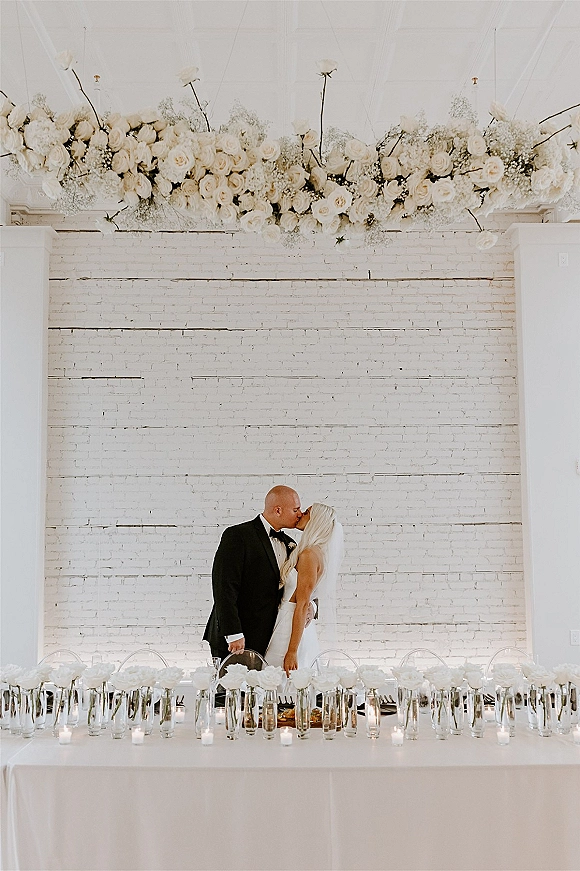 Wedding kiss portrait of bride and groom kiss at a candlelit head table with white roses and hanging flowers against a white brick wall