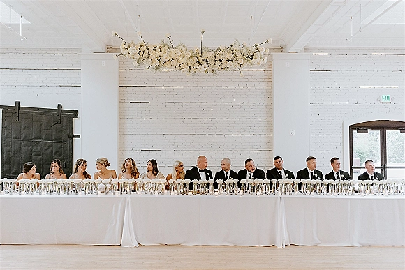 Wedding head table with bridal party seated, white floral garland and tall glass candles on a white tablecloth against a white brick wall