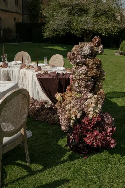Reception tablescape for an outdoor wedding table set with burgundy taper candles on a garden lawn beside a stone building.