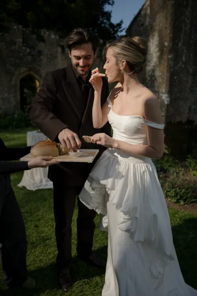 Reception candid of a couple tasting appetizers from a serving tray on a garden lawn near a stone arch and trees during cocktail hour.