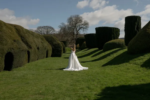 Bridal portrait of a bride in garden, showcasing the back of a low-back wedding dress with a sweeping dress train by topiary hedges.