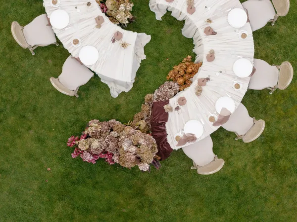 Reception tablescape in a serpentine table layout with a brown table runner, set on a grass lawn for an al fresco garden wedding.