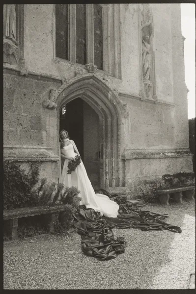 Bridal portrait of a bride with bouquet framed by a stone archway and ivy against a historic facade in a quiet gravel courtyard.