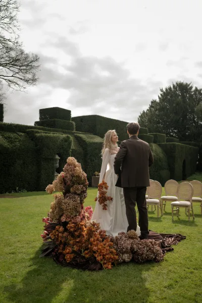 Ceremony moment at an outdoor garden ceremony as the bride and groom stand amid a sculptural floral installation, framed by topiary hedges.