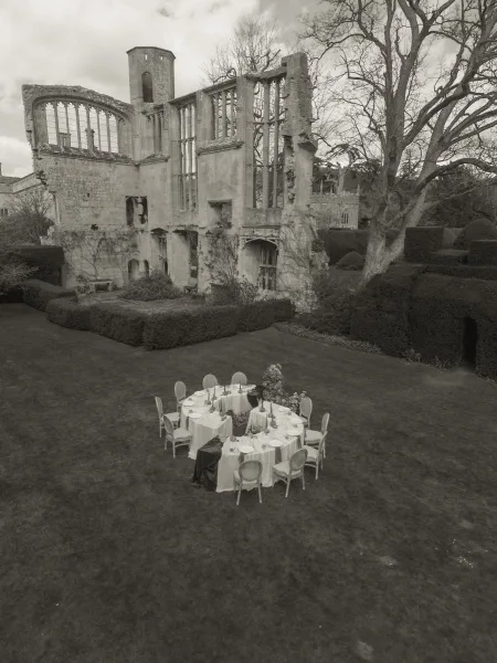 Round reception table for an outdoor reception setup with taper candles, on a lawn beside stone ruins under a cloudy sky.