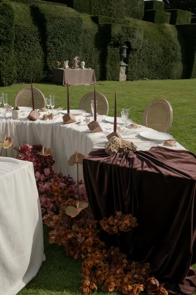 Reception tablescape styled for outdoor reception decor, featuring orchids on a garden lawn bordered by topiary hedges and a stone urn.