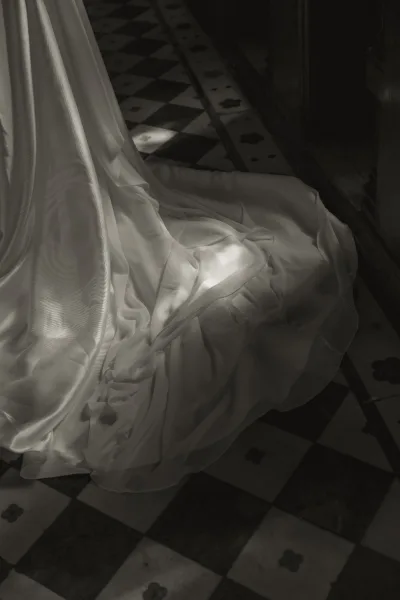 Wedding dress train in a bridal gown close-up, with soft fabric pleats pooling across a checkered tile floor and light on the fabric.