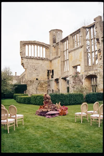 Ceremony setup with garden ceremony decor and grounded florals framing a non-arch altar against stone ruins and manicured hedges.