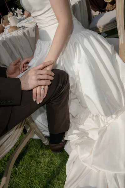 Couple holding hands, the bride and groom's hands display a diamond engagement ring at an outdoor reception table set on a grass lawn.