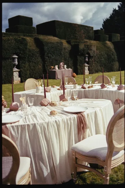 Reception tablescape for outdoor reception decor, featuring taper candles in a hedge garden with stone urns under a cloudy sky.