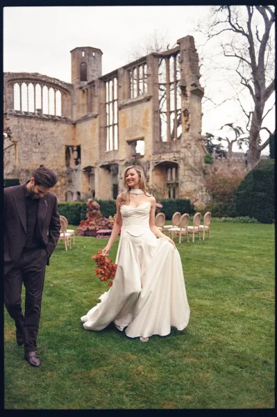 Couple portrait of the bride walking with bouquet beside her groom, strolling across a lawn with stone ruins and hedges behind them.