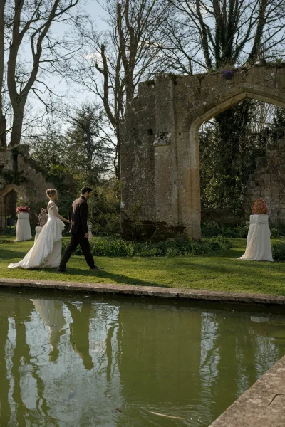 Bride and groom walking in a garden wedding portrait by stone ruins and an archway, passing floral arrangements beside a reflecting pond.