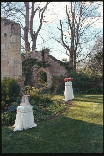 Ceremony decor featuring draped fabric by an arched doorway amid stone ruins, set on a garden lawn with trees and shrubs beyond.