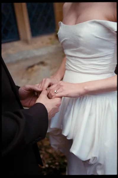 Ring exchange captured as an engagement ring close-up, the couple’s hands poised; bride in an off-shoulder gown by a stone wall and leaded windows.