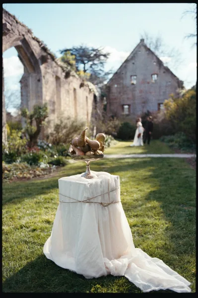 Pear centerpiece styled as fruit wedding decor in a silver compote bowl, set by stone ruins with a distant couple on the garden path.