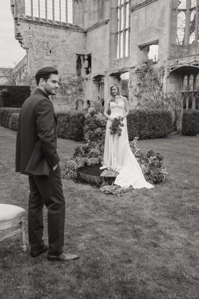 Couple portrait, a black and white wedding portrait where the bride holds a bouquet amid stone ruins with tall windows and garden hedges.