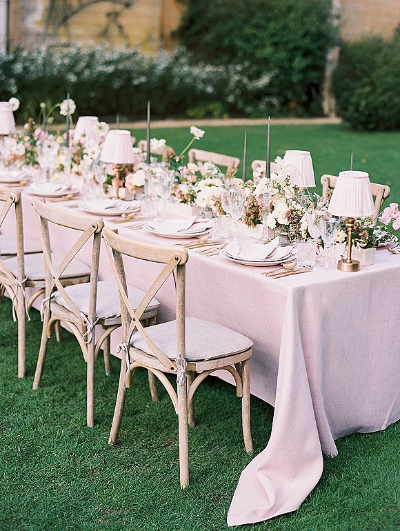 Reception tablescape with an outdoor reception table on a blush tablecloth, floral garland, taper candles, and pink table lamps on a garden lawn
