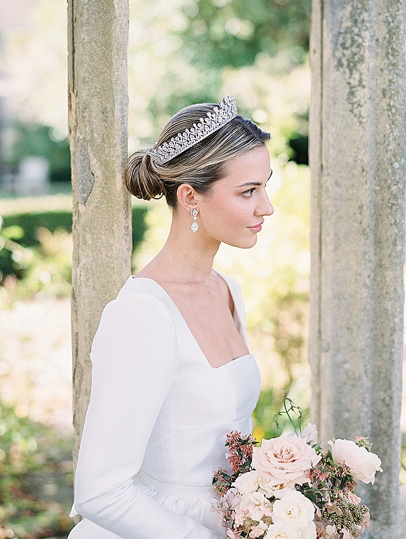 Bridal portrait with a crystal tiara, sleek low bun, and drop earrings, holding a blush rose bouquet beside stone columns and garden greenery