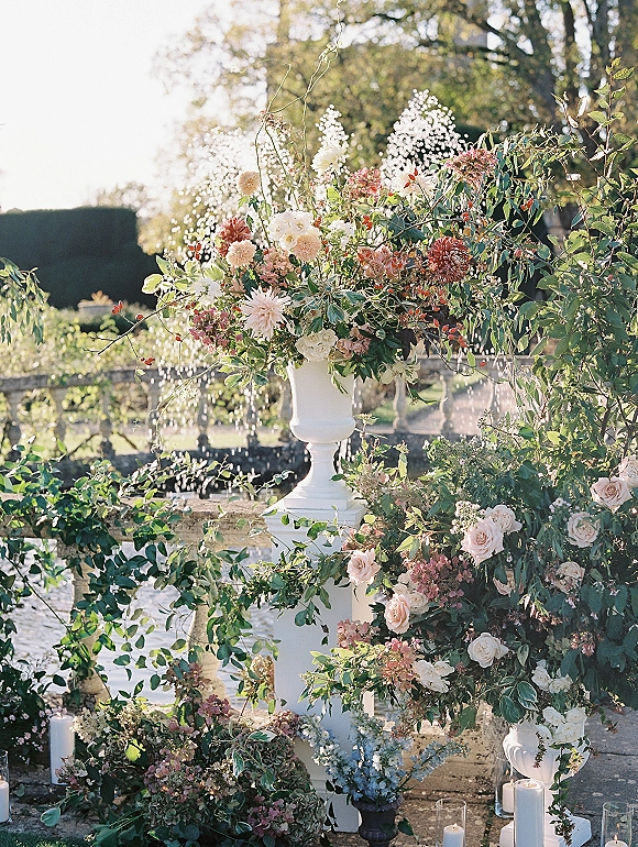 Ceremony floral arrangement in a white urn pedestal with roses, dahlias, baby’s breath and trailing greenery, set by stone balustrade candles