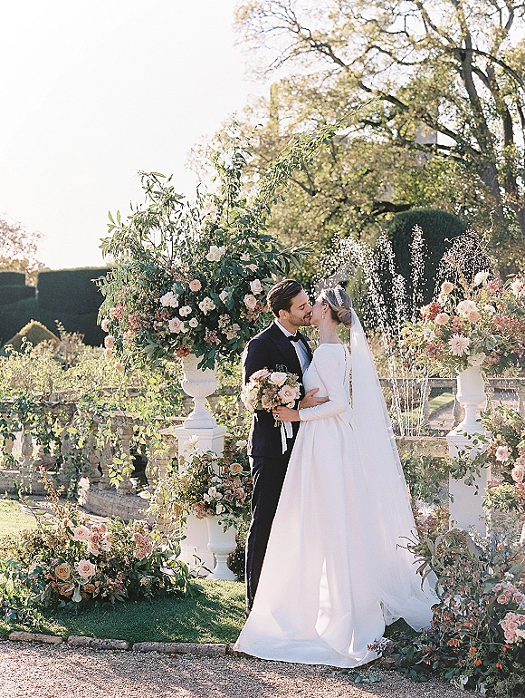 Wedding kiss portrait of bride and groom kissing by a fountain, her veil and bouquet visible beside floral urns and stone balustrade