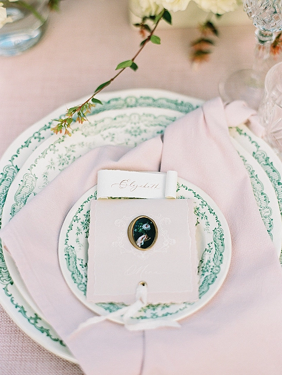 Wedding place setting with green and white plates, a pink linen napkin, ribbon-tied place card, crystal glassware on a linen tablecloth