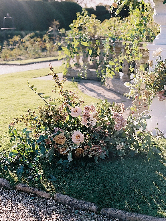 Ceremony floral arrangement of blush roses and greenery at the base of a floral arch beside a stone balustrade in sunlight garden lawn