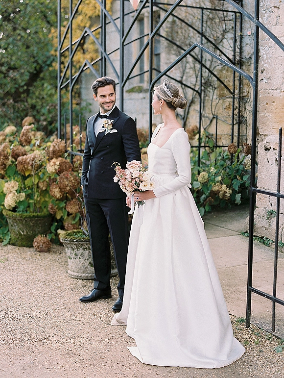 Couple portrait of bride and groom holding hands, groom gazing at bride with bouquet, beside hydrangeas and a stone wall trellis