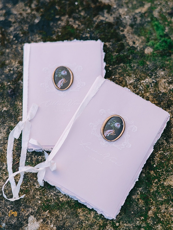 Invitation suite of deckled edge invitations wrapped in white ribbon with an oval wax seal, styled on a mossy stone surface