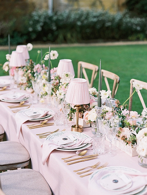 Reception tablescape with an outdoor reception table dressed in a pink tablecloth, gold flatware, candles, lamps, and floral centerpieces on a lawn