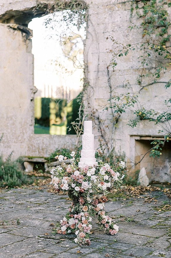 Wedding cake display with a tiered white wedding cake, lush floral arrangement and greenery set by a vine-covered stone ruin wall
