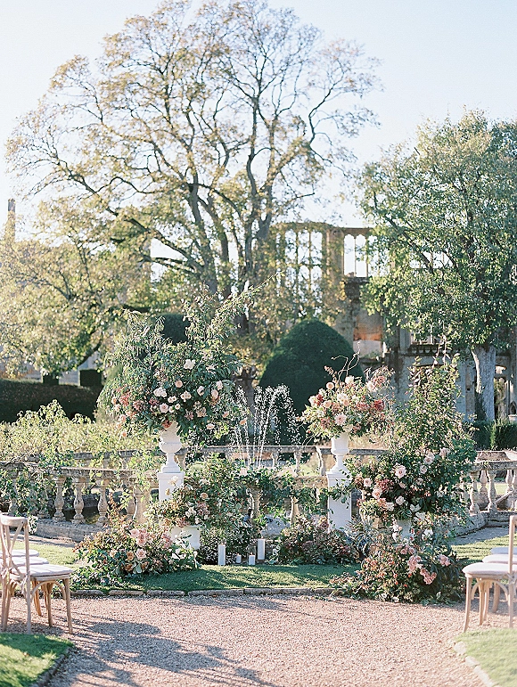 Ceremony setup for an outdoor garden ceremony with white chairs, gravel aisle, urn florals and candle accents near a fountain and stone balustrade