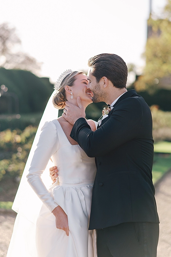 Wedding kiss portrait of bride and groom kissing as he cradles her face, her tiara and veil glowing in sunlit garden greenery