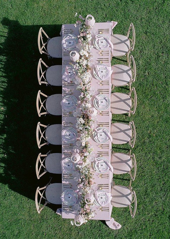 Reception tablescape with blush tablecloth, floral garland centerpiece, gold flatware, glass chargers, and candles on a green lawn outdoors