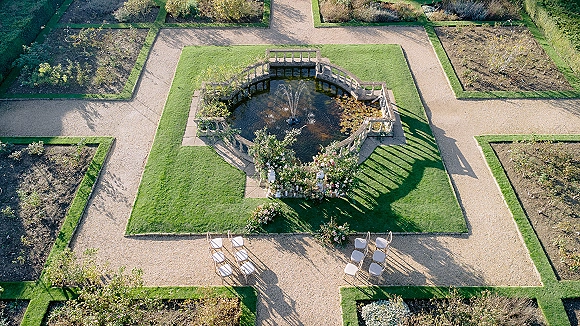 Outdoor ceremony setup with garden wedding ceremony chairs lining a gravel aisle, pastel florals and candle pedestals framing a fountain in a formal garden