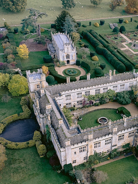 Wedding venue exterior with stone manor turrets and ivy, framing a circular fountain courtyard lawn, formal hedges, and sheep fields beyond