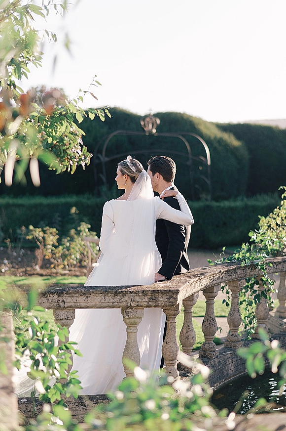 Couple portrait of bride and groom from behind, bride’s arm around him as they lean on a stone balustrade by a sunlit pond