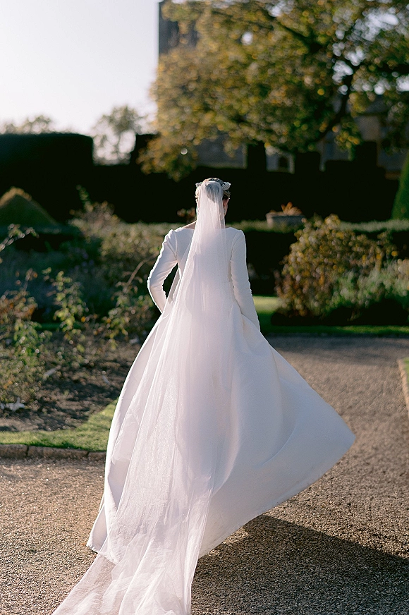 Bridal portrait of a bride walking away in a wedding dress with long veil and hair accessory on a sunlit garden path with hedges