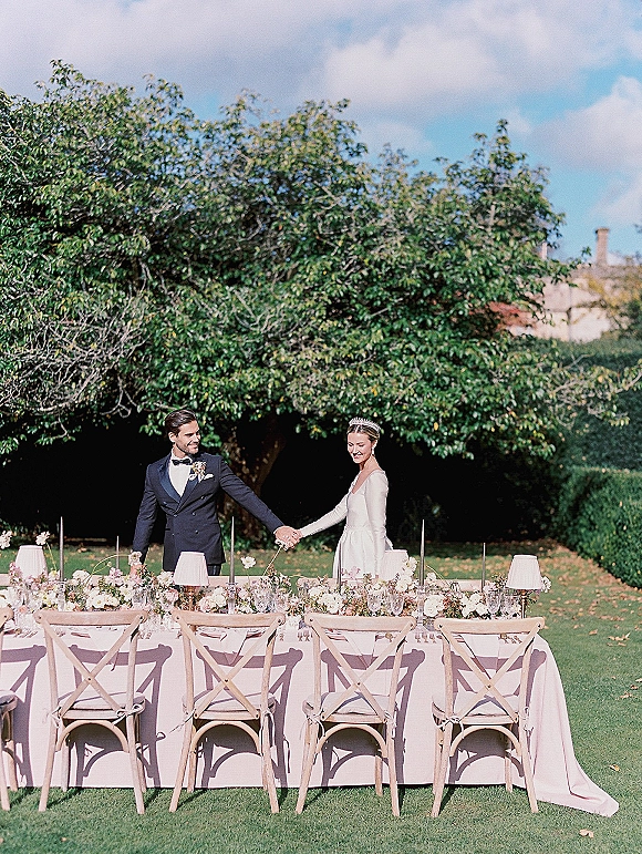 Newlywed couple holding hands behind a pink sweetheart table with taper candles and floral runner on a sunny garden lawn under trees
