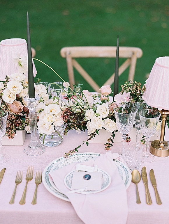 Reception tablescape with wedding table centerpiece of white roses and greenery garland, black taper candles, pink linens, and gold flatware on a green lawn