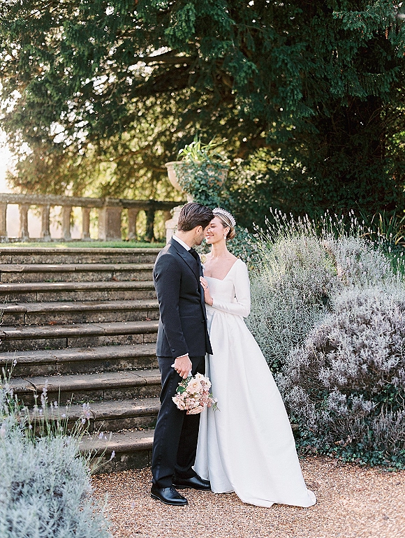 Couple portrait of bride and groom in tuxedo and long sleeve gown, holding a blush bouquet on stone steps amid garden greenery.