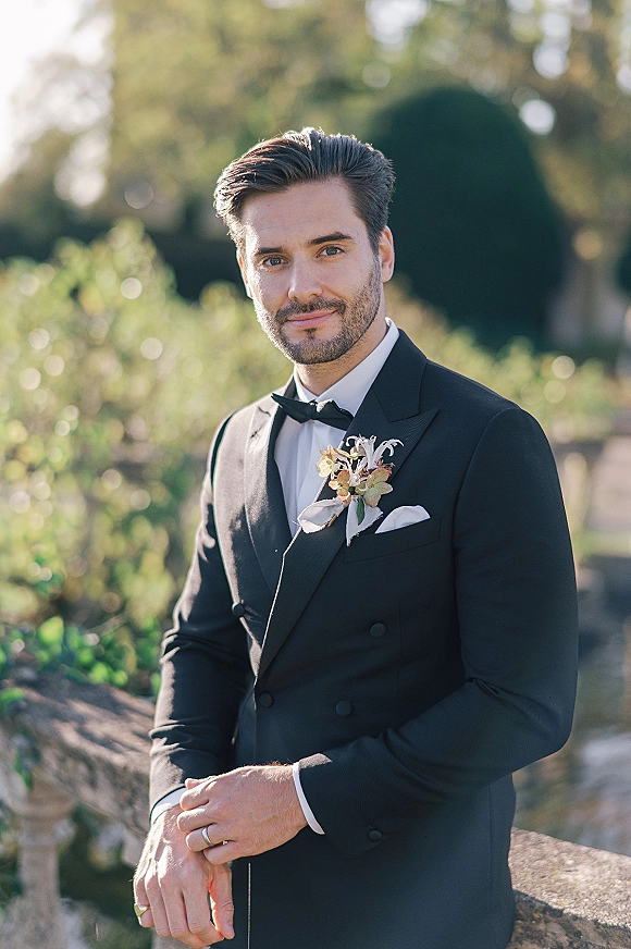 Groom portrait in a black tuxedo with bow tie, boutonniere, and wedding band, leaning on a stone railing in sunlit garden greenery.