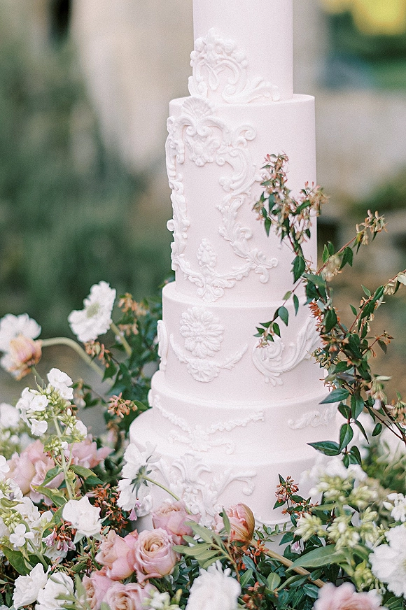 Wedding cake with ornate white scrollwork and floral appliques, topped with pink roses and greenery against a blurred garden backdrop