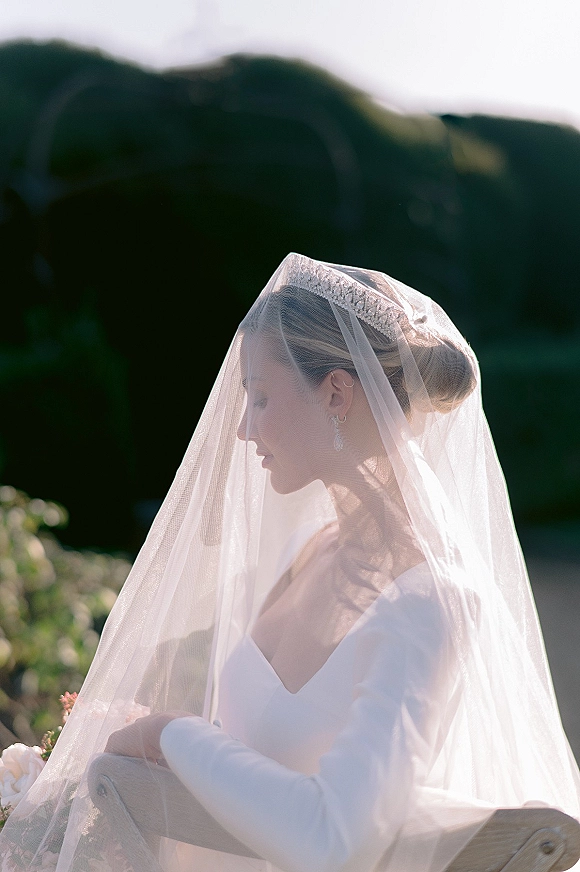 Bridal portrait of a bride wearing veil, seated with bouquet and tiara on a wooden chair, mountains and garden greenery behind her.