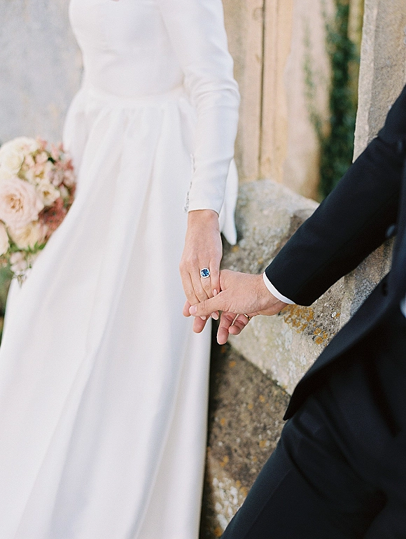 Wedding couple hands clasped with an engagement ring close up, bride’s long sleeve gown and blush bouquet against a stone wall backdrop