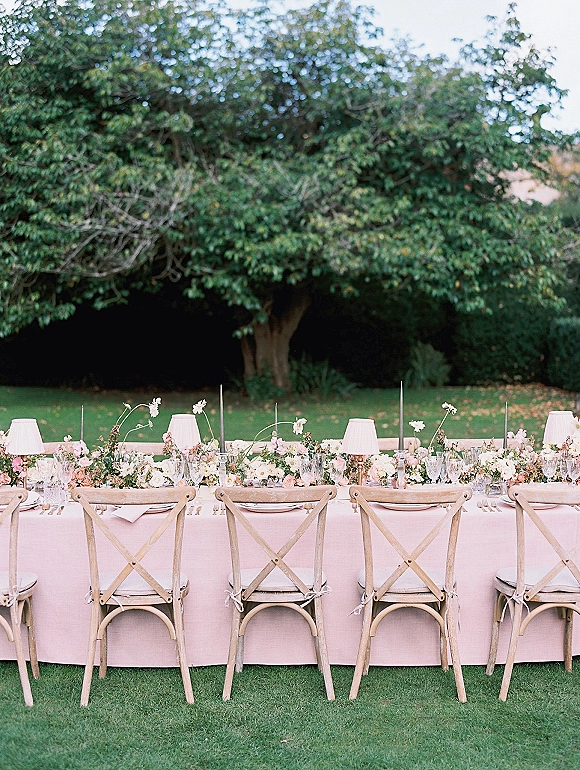 Reception tablescape with an outdoor wedding reception table on a long blush banquet setup, floral centerpieces, taper candles, and lawn greenery backdrop