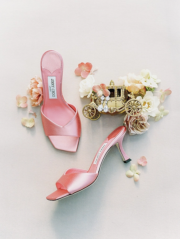 Bridal shoes flatlay featuring pink satin high heel mules with crystal drop earrings and soft petals on a light neutral backdrop